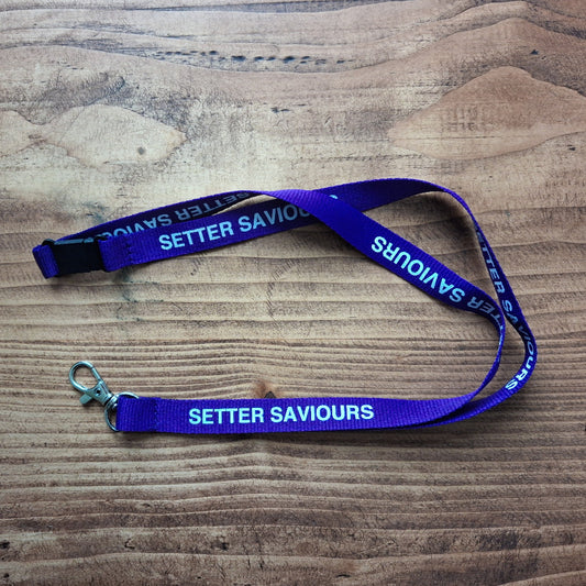 A purple lanyard with white text reading 'SETTER SAVOURS' laid out on a wooden surface, featuring a silver metal clasp and a black plastic connector.
