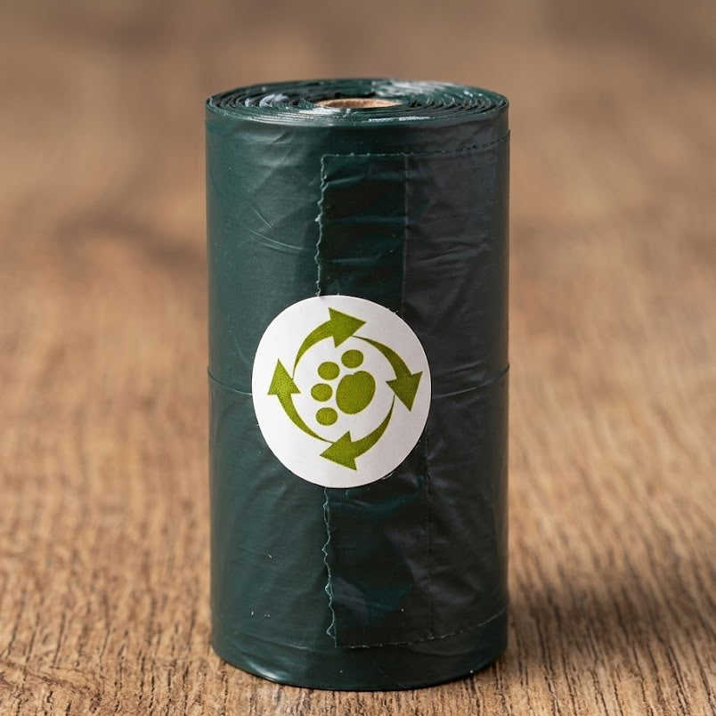 A roll of dark green biodegradable plastic dog poo bag standing upright on a wooden surface, featuring a white circular label with a green paw print and recycling arrows.