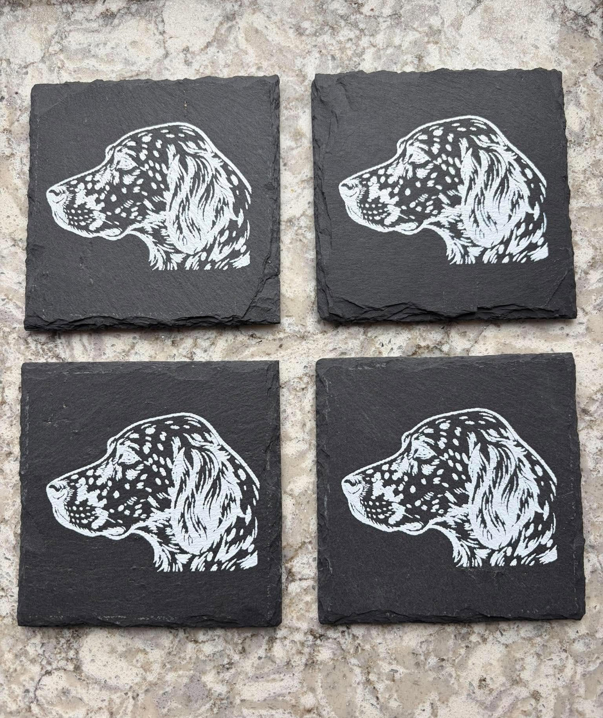 Four square black slate coasters arranged in a 2x2 grid on a light beige stone surface, each printed with a white dog head profile illustration.