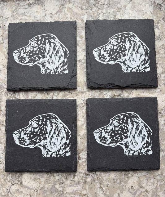 Four square black slate coasters arranged in a 2x2 grid on a light beige stone surface, each printed with a white dog head profile illustration.